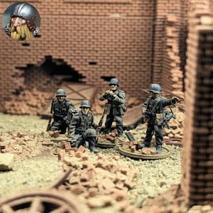 Peut inclure: Figurines miniatures de soldats en uniformes et casques gris, posées dans un environnement ravagé par la guerre avec des ruines de briques. Une figurine est agenouillée, tandis que d'autres sont debout, tenant des fusils et de l'équipement. La scène se déroule sur une surface sablonneuse avec des débris éparpillés.