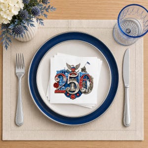 Puede incluir: Una mesa puesta con un plato blanco con borde azul, un tenedor y un cuchillo. Una pila de servilletas blancas con un dise&ntilde;o patri&oacute;tico con el n&uacute;mero 250, un &aacute;guila y otros s&iacute;mbolos patri&oacute;ticos. Un vaso azul y un jarr&oacute;n de flores completan el conjunto.