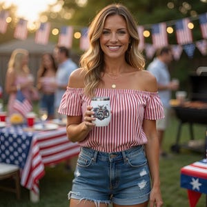 Op de afbeelding: Een witte roestvrijstalen tumbler met een motorfietsafbeelding. De tumbler wordt vastgehouden door een persoon die een rood-wit gestreepte off-the-shoulder top en een spijkerbroek draagt. De achtergrond bevat Amerikaanse vlaggen en een barbecue.