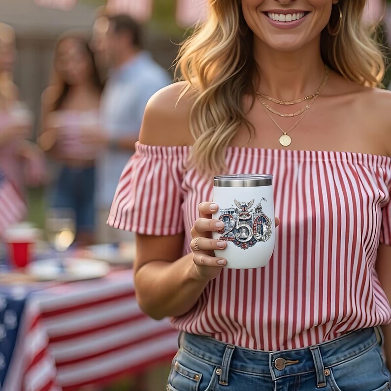 Op de afbeelding: Een witte ge&iuml;soleerde tumbler met een zilveren rand, met een patriottisch ontwerp en het nummer "250". De tumbler wordt vastgehouden door een persoon die een rood-wit gestreept off-the-shoulder top en een spijkerbroek draagt. De achtergrond suggereert een buitenevenement.