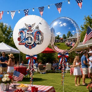 May include: Two round balloons, one white with a patriotic design celebrating 250 years, and one reflective silver, are displayed outdoors. The white balloon features an eagle, the Liberty Bell, and the numbers 250 and 1776-2026. Both balloons have red, white, and blue ribbons.
