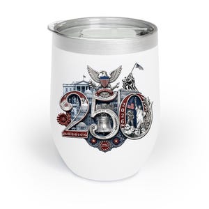 Op de afbeelding: Witte ge&iuml;soleerde wijnbeker met een zilveren rand en deksel. De beker heeft een patriottisch ontwerp met de tekst "250 AMERICA 1776-2026" en illustraties van het Amerikaanse Capitool, de Liberty Bell en het Vrijheidsbeeld.