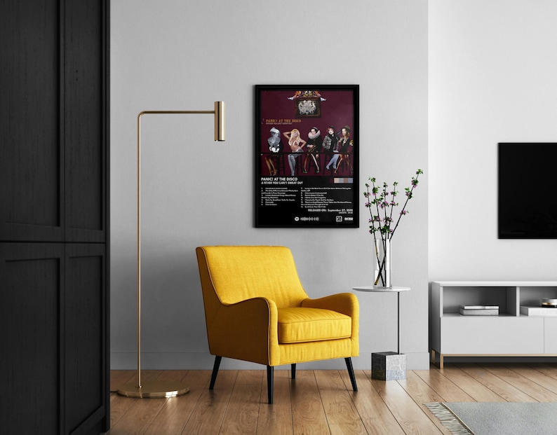 Op de afbeelding: Een zwart ingelijste poster met een bandfoto en de tekst "Panic at the Disco" en "A Fever You Can't Sweat Out". De poster hangt aan een witte muur boven een gele fauteuil.