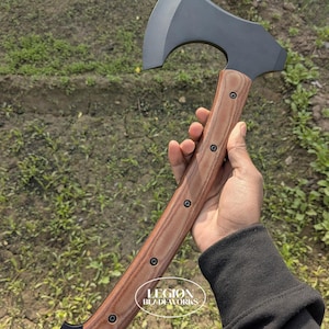 May include: Axe with a dark gray blade and a wooden handle. The handle is brown with black accents and is secured with black rivets. The axe is held in a hand, with the text "LEGION BLADEWORKS" visible on the handle.