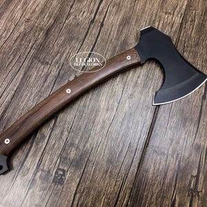 Puede incluir: Un hacha con cabeza de metal negro mate y mango de madera marrón oscuro curvado, asegurado con tres remaches plateados. La cabeza presenta un filo plateado pulido. El texto LEGION BLADEWORKS está impreso en blanco sobre el mango.