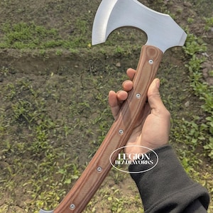 Op de afbeelding: Een handbijl met een zilverkleurige metalen kop met een gebogen blad en een lange, bruine, getextureerde houten steel die is vastgezet met vijf zilveren klinknagels. De onderkant van de steel heeft een zilveren metalen dop met twee gaten. De tekst LEGION BLADEWORKS is zichtbaar.