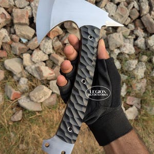 May include: A metal hatchet with a polished silver blade and a textured black handle featuring a repeating scalloped pattern. The handle is secured with three silver rivets and includes two circular holes at the base. The text LEGION BLADEWORKS is visible.