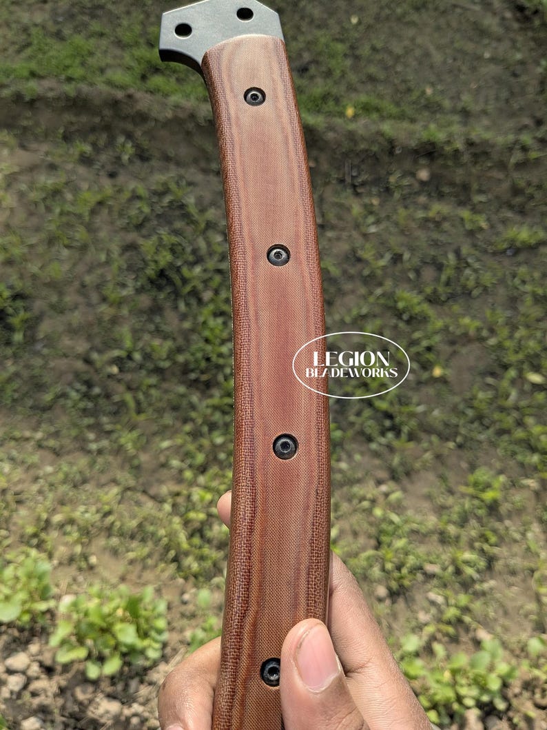 May include: Close-up of a knife handle with a brown textured grip and black screws. The blade's metal top is visible. The text "LEGION BLADEWORKS" is printed on the handle.