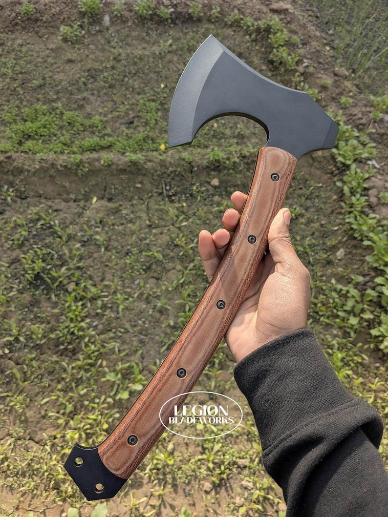 May include: Axe with a dark gray blade and a wooden handle. The handle is brown with black accents and is secured with black rivets. The axe is held in a hand, with the text "LEGION BLADEWORKS" visible on the handle.