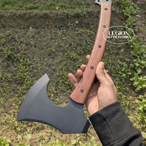 May include: Axe with a dark gray blade and a curved wooden handle. The handle is brown with visible wood grain and is secured with rivets. The axe head has a curved cutting edge. The text "LEGION BLADEWORKS" is visible on the handle.