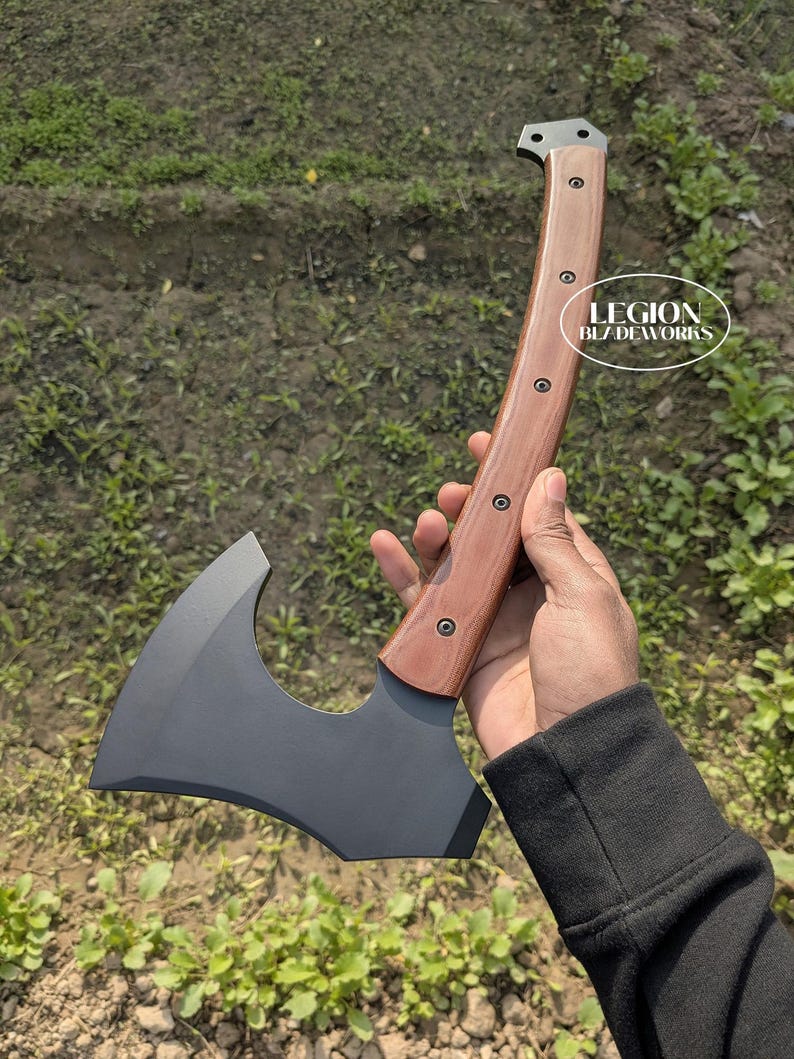 May include: Axe with a dark gray blade and a curved wooden handle. The handle is brown with visible wood grain and is secured with rivets. The axe head has a curved cutting edge. The text "LEGION BLADEWORKS" is visible on the handle.