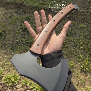May include: Axe with a dark gray blade and a wooden handle. The handle is light brown with dark rivets. The axe has a curved blade and a hammer on the back. The text "LEGION BLADEWORKS" is in a white oval.
