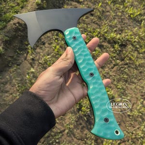 Op de afbeelding: Een zwarte tomahawk met een turquoise getextureerde handgreep. Het mes heeft een unieke vorm met een puntige rand. De handgreep is vastgezet met drie zwarte klinknagels. Het logo "LEGION BLADEWORKS" is zichtbaar.