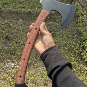 May include: A dark gray tomahawk axe with a wooden handle. The handle is a light brown color with dark brown rivets. The axe head is a dark gray color. The axe is held in a hand.