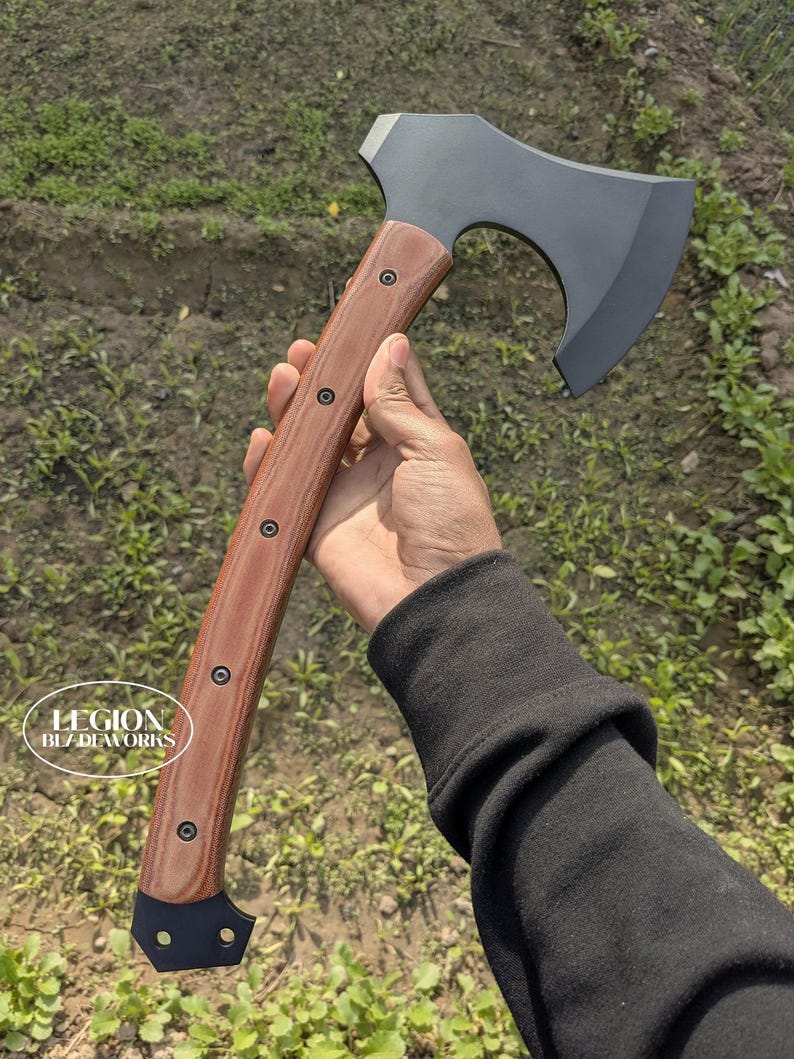 May include: A dark gray tomahawk axe with a wooden handle. The handle is a light brown color with dark brown rivets. The axe head is a dark gray color. The axe is held in a hand.