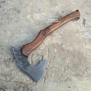 Op de afbeelding: Een handbijl met een donkere, getextureerde, gesmede metalen kop en een gebogen, gepolijste bruine houten steel met zichtbare houtnerf. De steel is bevestigd met metalen klinknagels. De tekst LEGION BLADEWORKS staat in het wit op de steel.