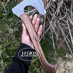 Może przedstawiać: Top&oacute;r z ciemnym drewnianym trzonkiem i szarym metalowym ostrzem. Rękojeść jest nitowana małymi srebrnymi szpilkami. Tekst "LEGION BLADEWORKS" znajduje się w białym owalu. Tekst "SEIRRA SIX" jest w białych literach blokowych.