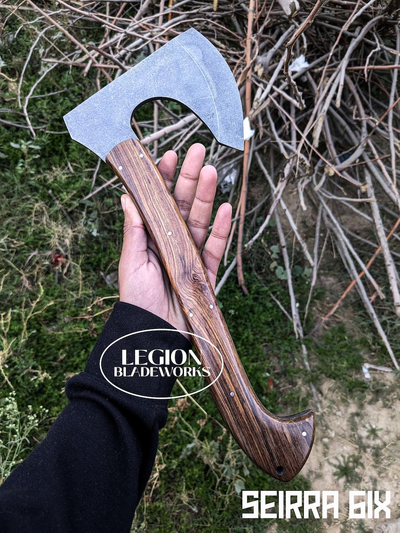 Może przedstawiać: Top&oacute;r z ciemnym drewnianym trzonkiem i szarym metalowym ostrzem. Rękojeść jest nitowana małymi srebrnymi szpilkami. Tekst "LEGION BLADEWORKS" znajduje się w białym owalu. Tekst "SEIRRA SIX" jest w białych literach blokowych.