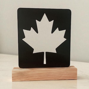 Puede incluir: Un recorte de hoja de arce de metal negro sobre una base de madera. La hoja de arce es un símbolo nacional de Canadá. La base de madera es rectangular y tiene un acabado de vetas de madera natural. El fondo es de color neutro.