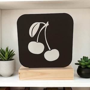 May include: A black square sign with rounded corners featuring a white cherry design. The sign is mounted on a natural wood base and displayed on a white shelf with books and potted plants.