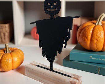 Könnte beinhalten: Schwarze Halloween-Dekoration eines Vogelscheuche mit geschnitztem Kürbiskopf und zerfetztem Körper. Die Vogelscheuche ist auf einem hellen Holzsockel montiert. Zwei orangefarbene Kürbisse befinden sich auf beiden Seiten.