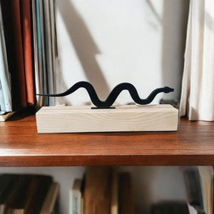 May include: A black metal snake sculpture on a light wood base. The sculpture is on a shelf with books behind it.