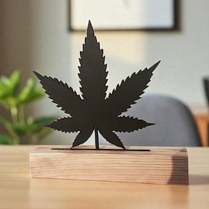 Peut inclure: Sculpture en métal noir en forme de feuille de cannabis, posée sur un bloc de bois clair. La sculpture est exposée sur un bureau en bois, avec une impression botanique encadrée en arrière-plan. Le bureau est devant une fenêtre et une chaise grise.