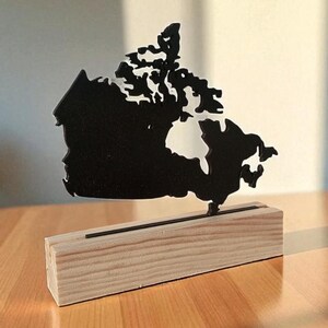 Peut inclure: Silhouette noire en métal représentant la carte du Canada, montée sur une base en bois naturel. La carte est une représentation détaillée du pays. La base en bois rectangulaire présente un grain de bois clair et naturel. L'ensemble est posé sur une table en bois.
