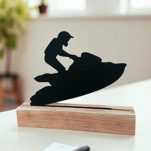 May include: A black silhouette of a person riding a jet ski, mounted on a rectangular wooden base. The jet ski rider is wearing a helmet. The wooden base is light brown. The background is a blurred interior scene.