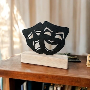 May include: Black wooden sculpture of two theater masks, one smiling and one frowning, on a wooden base. The masks are a traditional symbol of comedy and tragedy.