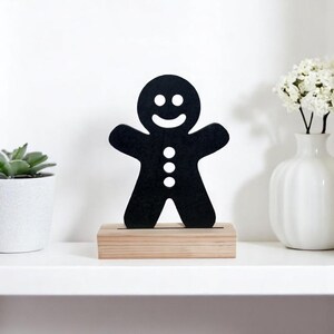 May include: Black gingerbread man cutout on a wooden stand. The gingerbread man has a smiling face and three buttons.