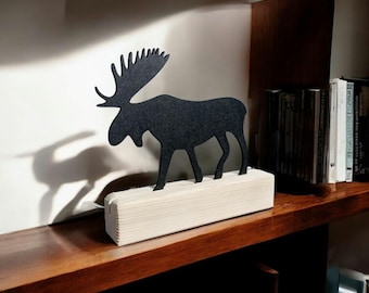 May include: A black moose silhouette on a wooden base, standing on a wooden shelf with books. The moose is facing left and has a long neck and antlers.