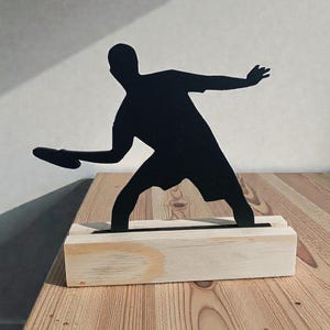 May include: Black silhouette of a person throwing a disc, mounted on a light-colored wooden base. The figure is in mid-throw, with one arm extended and the disc visible. The base is rectangular and appears to be made of unfinished wood.
