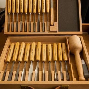 May include: A set of wood carving tools, including chisels and a mallet, presented in an open wooden box. The chisels have wooden handles and metal blades. The box is open, displaying the tools. The set also includes a knife and a small container.