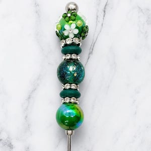 May include: A decorative pen featuring a series of green beads, including a top bead adorned with small white and green flowers, a glittery dark green bead, and a smooth metallic green bead, separated by silver-toned rhinestone spacers.