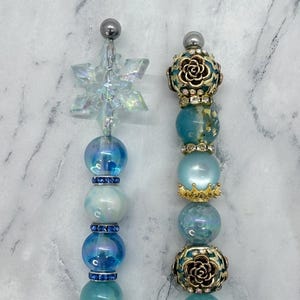 May include: Two decorative pens featuring assorted beads in shades of blue, teal, and white. The left pen is topped with a clear iridescent snowflake, while the right pen features metallic rose-patterned beads and gold-toned accents.
