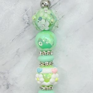 May include: A decorative pen topper featuring a vertical arrangement of mint green, white, and pastel-colored beads. The design includes floral-shaped beads, small heart accents, and silver-toned rhinestone spacers, topped with a metallic sphere.