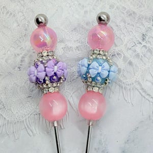May include: Two decorative pens featuring stacked beads with metallic silver accents. One pen has purple bow-shaped beads, while the other has light blue bow-shaped beads. Both pens are topped with a large, iridescent pink spherical bead.