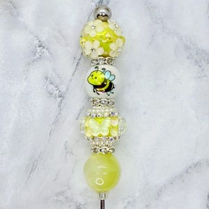 May include: A decorative pen topper featuring a stack of beads including a yellow bead with white flowers, a white bead with a cartoon bee, a clear bead with a yellow butterfly, and a smooth yellow bead, all separated by silver-toned rhinestone spacers.