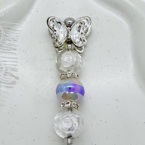 May include: A decorative pen topper featuring a silver-toned metal butterfly with clear crystal wings at the top. Below the butterfly are two clear beads with embossed rose patterns, separated by a central iridescent bead and two small rhinestone spacers.