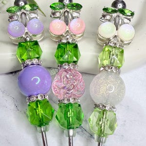 May include: Three decorative pens featuring silver-tone metal shafts topped with ornate beadwork. Each pen includes green crystal-like accents, rhinestone spacers, and distinct focal beads in lavender, pink, and white glitter finishes.