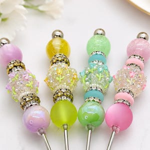 May include: Four decorative pens featuring beaded tops with various pastel colors including lavender, yellow, mint green, and pink. Each pen includes clear, glittery beads with floral patterns, metallic spacers with rhinestones, and smooth round beads.
