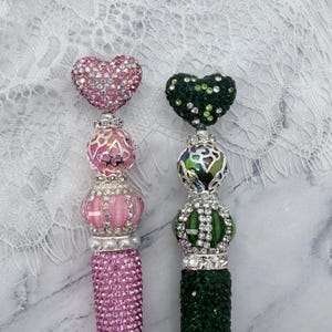 May include: Two decorative beaded tassels. One is pink with a heart-shaped top covered in pink rhinestones. The other is green with a heart-shaped top covered in green rhinestones. Both have ornate silver accents.