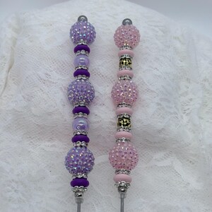 May include: Two beaded pens. One pen features purple beads and accents, the other has pink beads and accents. Both pens have silver-colored metal tips and tops. The pens are embellished with rhinestones and other decorative elements.