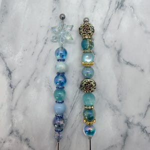 May include: Two decorative hat pins. One features a clear snowflake, blue beads, and a silver pin. The other has gold rose accents, blue and iridescent beads, and a silver pin. Both are elegant accessories.