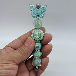 May include: A decorative teal butterfly bottle stopper. The stopper features a teal butterfly, a silver bead, and a series of teal beads with floral and crystal accents. The stopper is held in a hand against a white background.