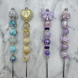 May include: Four decorative skewers with beaded designs. The skewers feature various colors, including blue, gold, and purple. Each skewer has a different top design, such as a heart, bow, and floral accents. The beads are of different sizes and shapes.