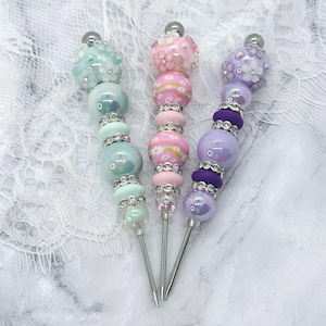 May include: Three decorative pens featuring stacked beads in pastel shades of mint green, pink, and purple. Each pen is topped with a floral-patterned bead and accented with silver-toned rhinestone spacers between the smooth, round, and patterned beads.