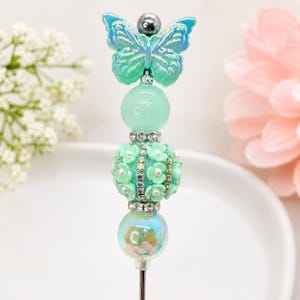 May include: A decorative pen topper featuring a light green iridescent butterfly at the top, followed by a smooth mint green bead, a textured mint green floral bead with small rhinestones, and a translucent iridescent bead at the base.