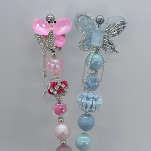 May include: Two decorative earrings featuring butterfly designs. One earring has a pink butterfly with silver accents and dangling beads. The other earring has a blue butterfly with silver accents and dangling beads. The earrings have a variety of pink and blue beads.